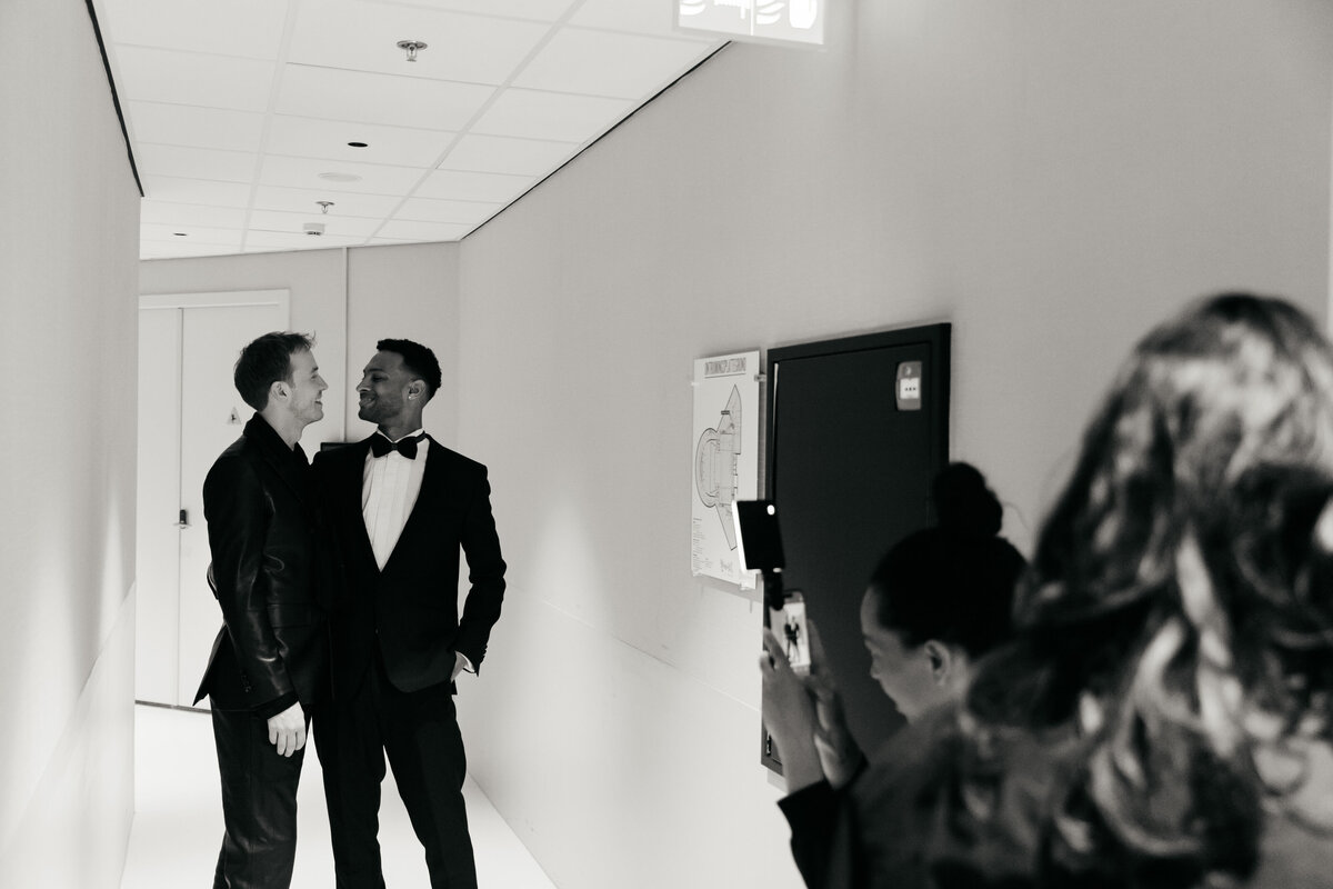 Twee mannen staan in een witte gang en lachen naar elkaar terwijl ze gefotografeerd worden door meerdere mensen.