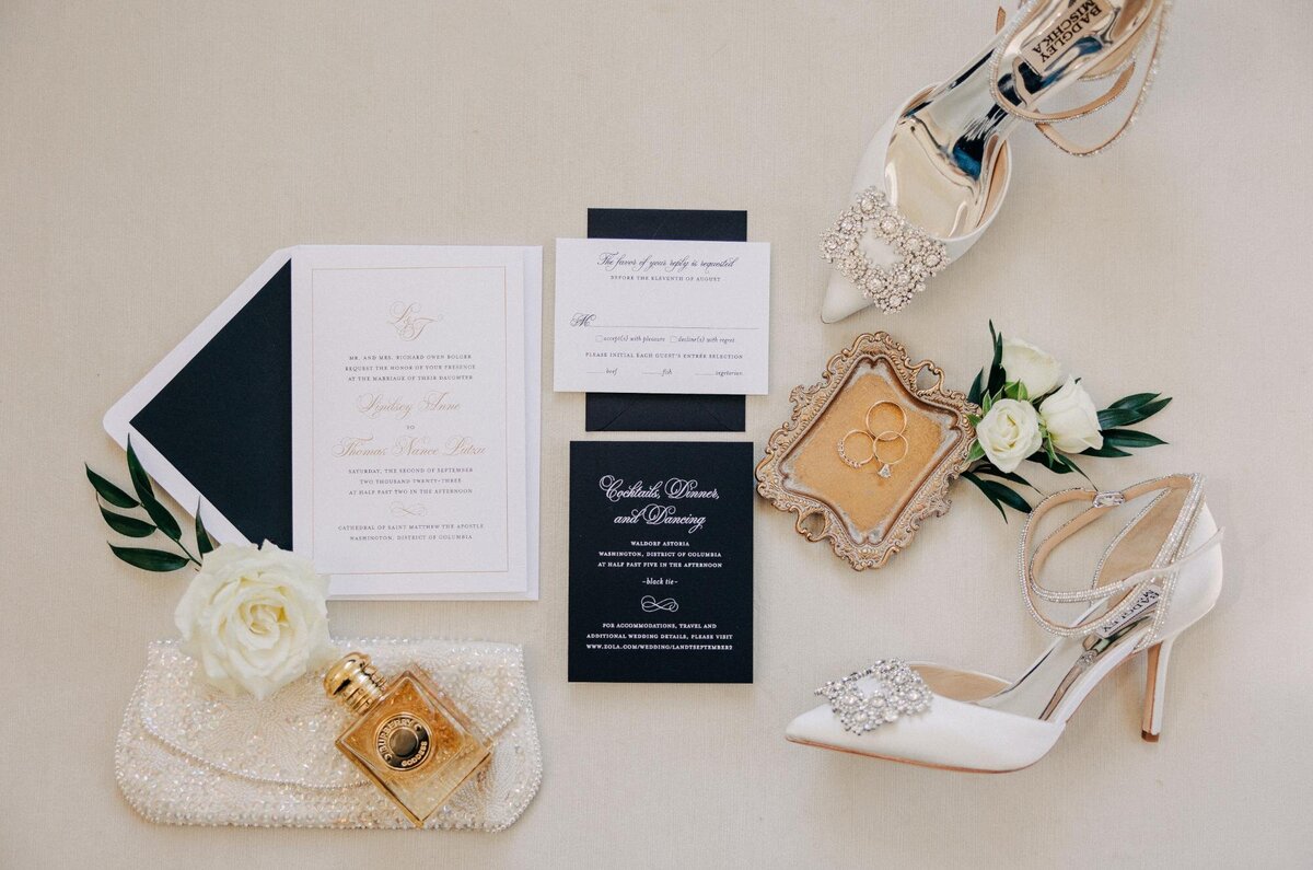 A wedding arrangement featuring invitations, a pair of white high-heeled shoes, a boutonniere, perfume, and a clutch on a neutral background.