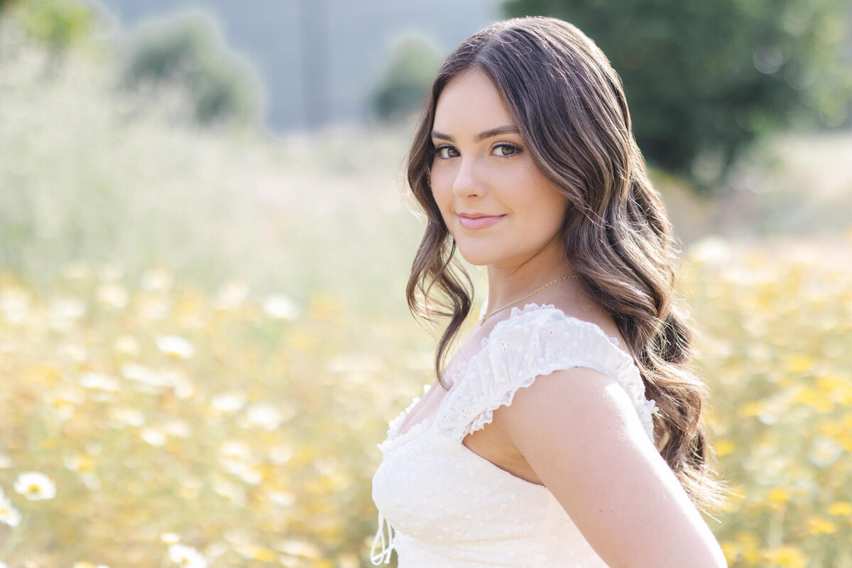 A close-up of a young woman with long, brown, wavy hair looking over her left shoulder at the camera, wearing a white dress, standing in tall green grass at Northwest Open Space in San Juan Capistrano.