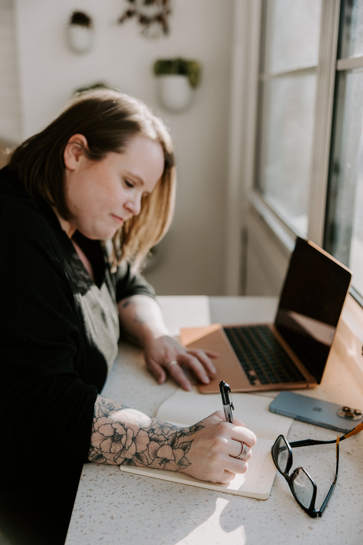 Creative female entrepreneur with floral tattoos writing business notes in a notebook at a bright, modern desk with laptop, smartphone, and glasses