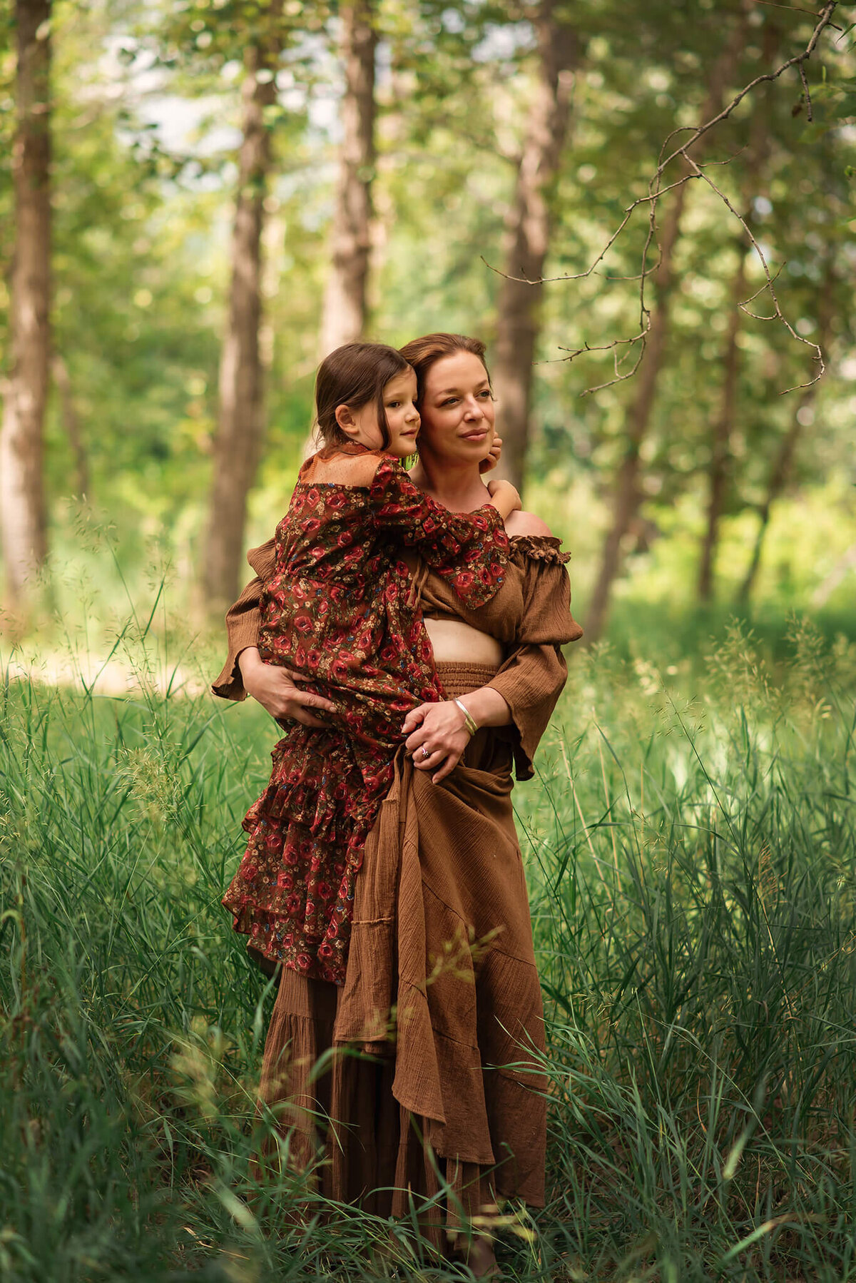 A mother and daughter stand in the tall green grass. Mother holds her young daughter on her hip and they book look off into the distance.  Portrait by Brittany Danielle
