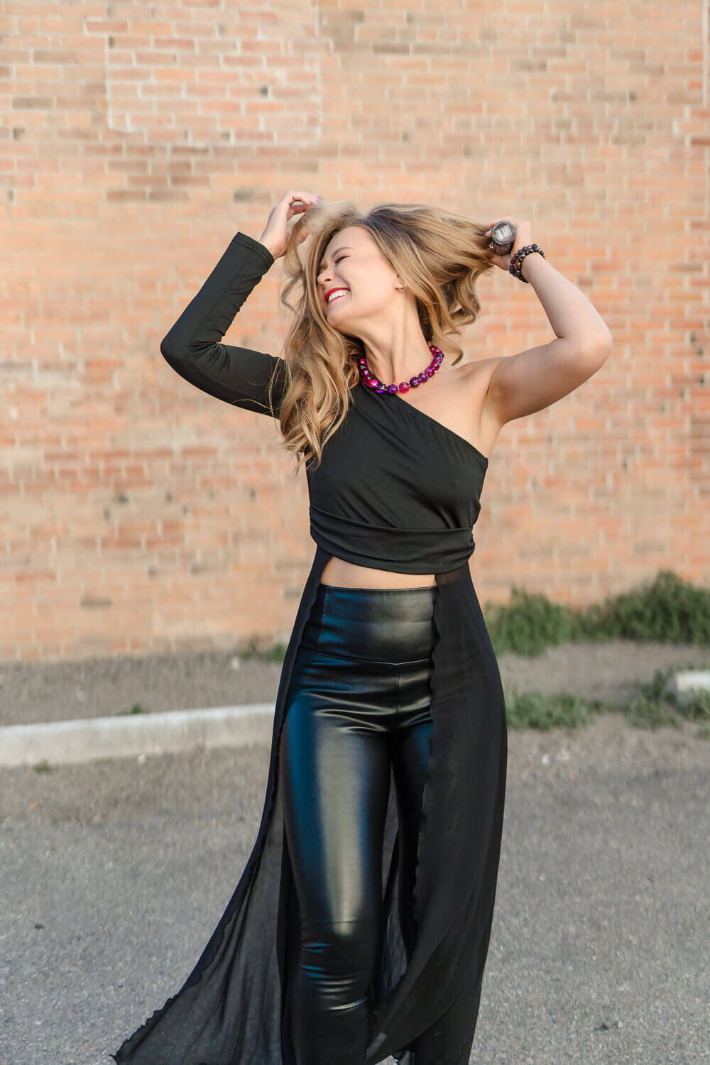 Entrepreneur in black leather pants and top posing confidently against a brick wall outdoors in the Okanagan.