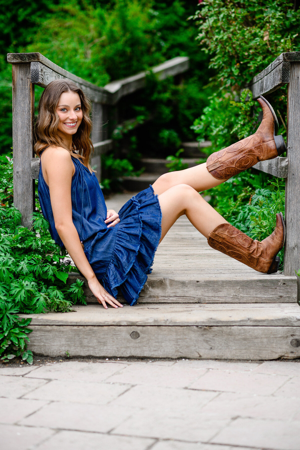 A girl in a jean dress and cowboy boots sitting on a bridge at the Denver Botanic Gardens for her Denver Senior Pictures