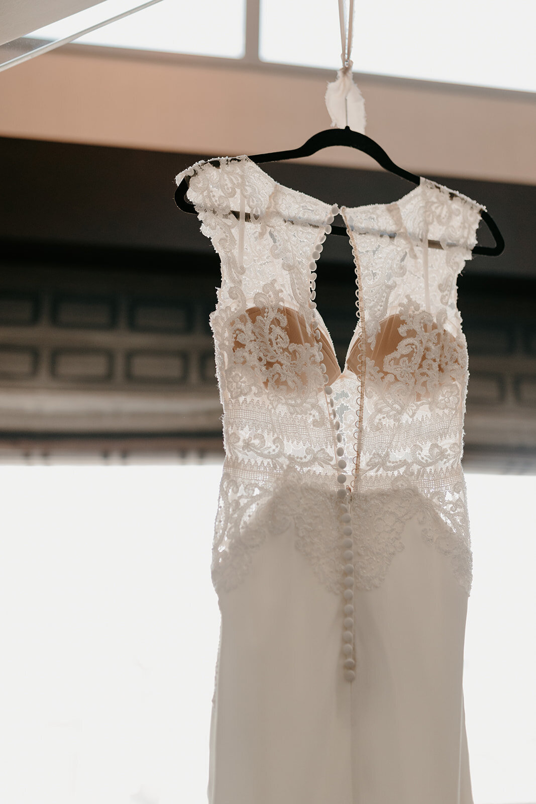 Close-up of the intricate lace details on the back of a white wedding dress with a row of buttons and delicate floral embroidery, hanging on a black hanger.