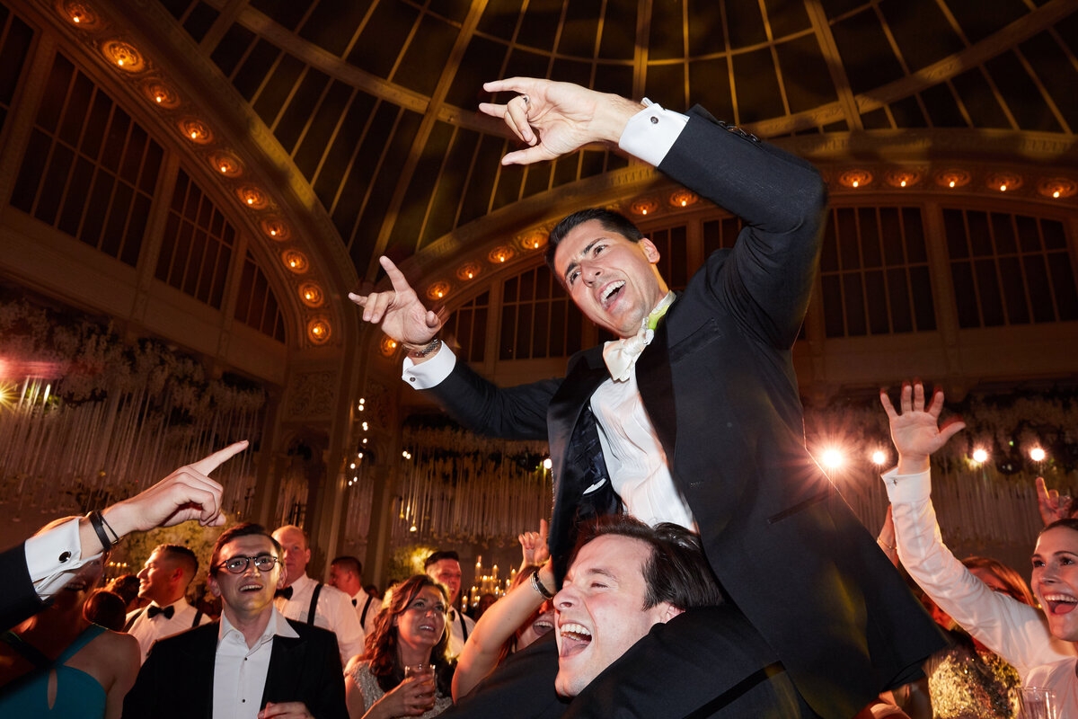 The event captured in the photo took place at The New York Public Library in New York City, beautifully documented by photographer Christian Oth and videographers East West Films. Within the opulent setting of the venue, a group of individuals dressed in formal attire are joyously celebrating. Among them, a man in a stylish tuxedo is being lifted up by another gentleman, his smiling face and exuberant hand gestures capturing the essence of the festive moment. The atmosphere is filled with excitement, as the onlookers surrounding them cheer and applaud in delight.
