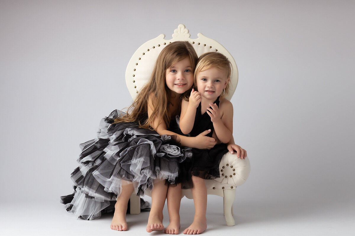 Two sisters smiling together in matching black and white dresses, sitting close in a vintage chair.