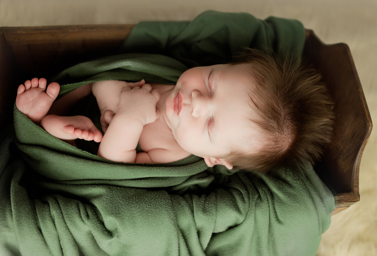 Newborn baby boy, laying in wood box wrapped in green wrap during newborn photoshoot in Mount Juliet Tennessee photography studio