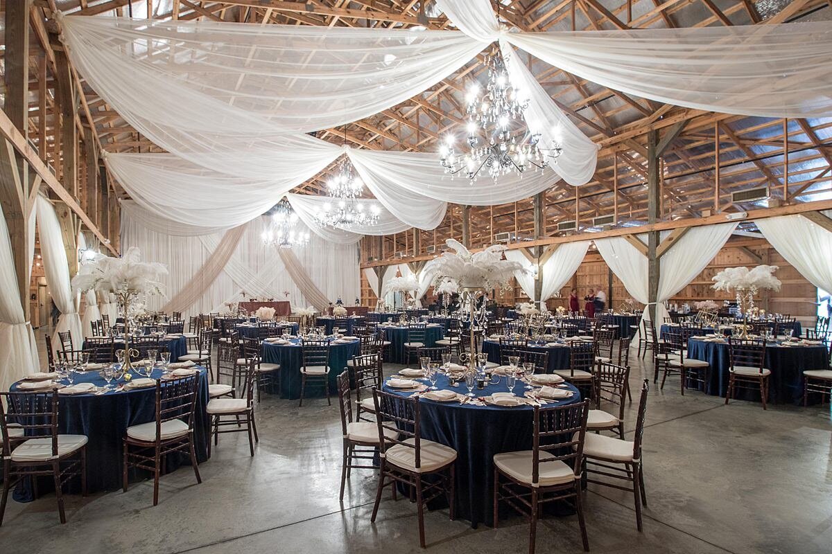 A 1920s themed wedding reception at Saddle Woods Farm decorated with navy blue velvet table cloths, brown chiavari chairs with ivory cushions tall crystal and ostrich feather centerpieces and a lot of sheer ivory drapery. The ceiling is draped with sheer champagne and ivory fabric accenting the large crystal and gold candelabra chandeliers of the cedar wood reception barn at Saddle Woods Farm
