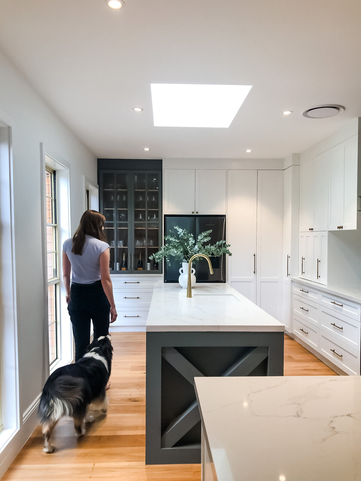 A woman strolls with her dog through a modern kitchen, boasting a large island and skylight, crafted by a bespoke interior design team from Newcastle NSW.