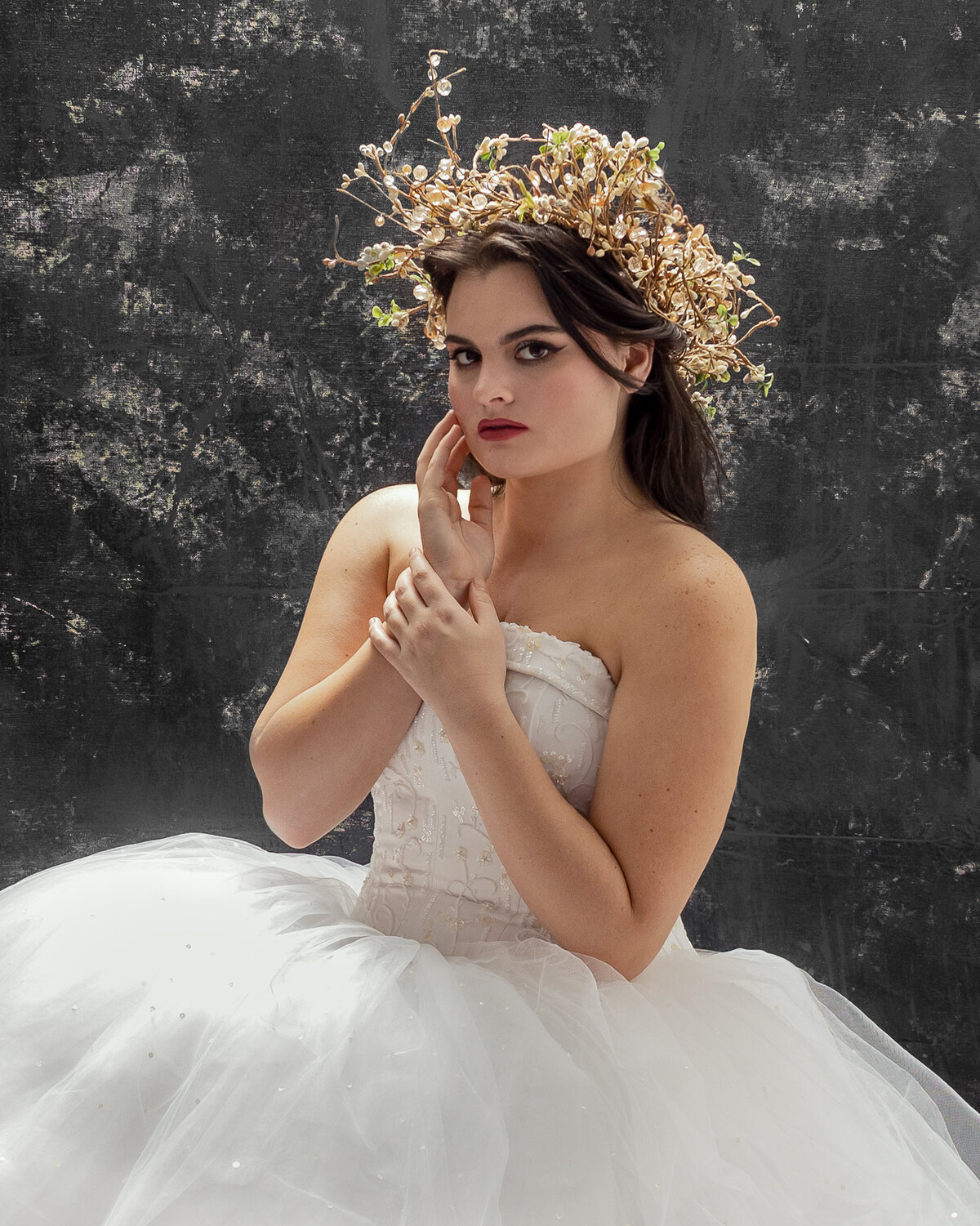 woman in wedding dress and a crown of flowers holding right wrist and looking  forward with right hand on face
