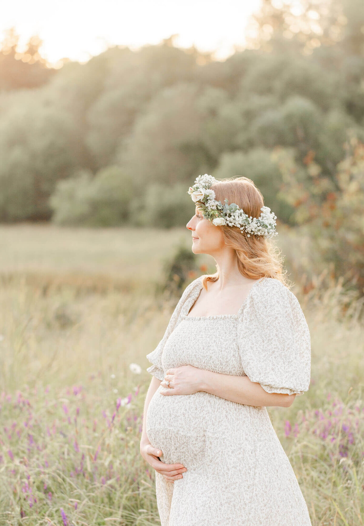 Pregnant woman in a nature field with wildflowers at sunset. She is holding her belly, looking out at the sunset wearing a floral crown during her Portland Oregon sunset maternity photo session.