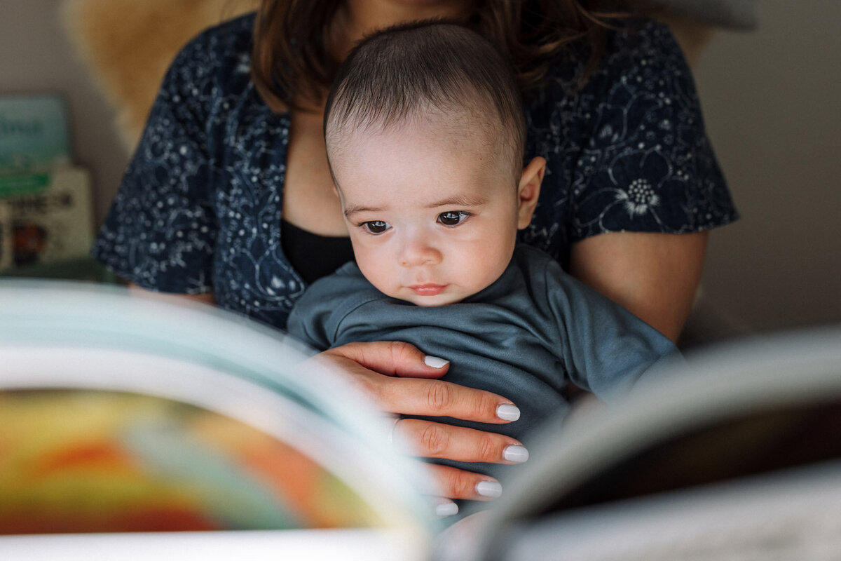 baby sitting in mom's lap as she gently touches his belly and they look at a book together