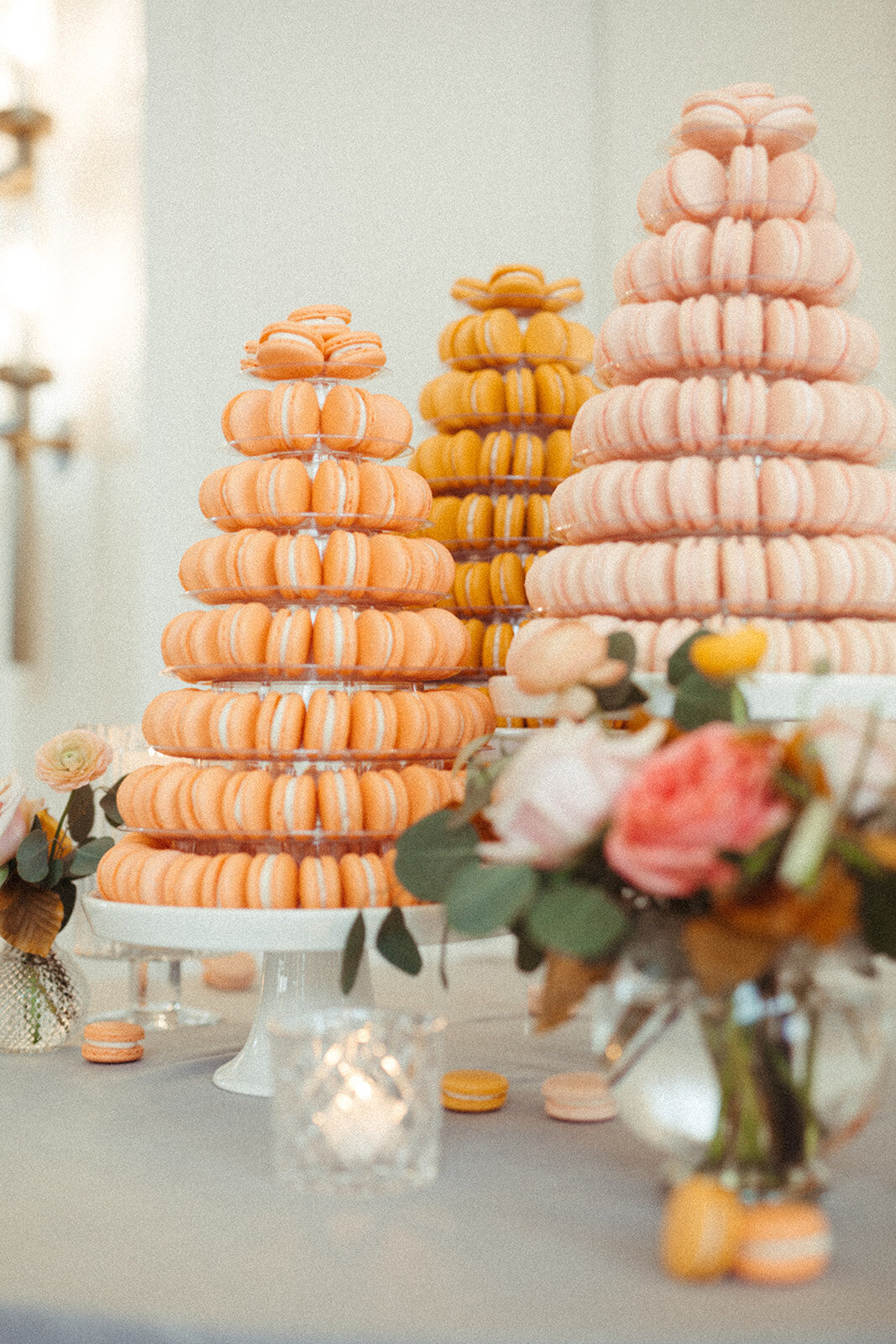 An assortment of macaroons on cake stands atop a table with light gray linen, a candle and flowers.