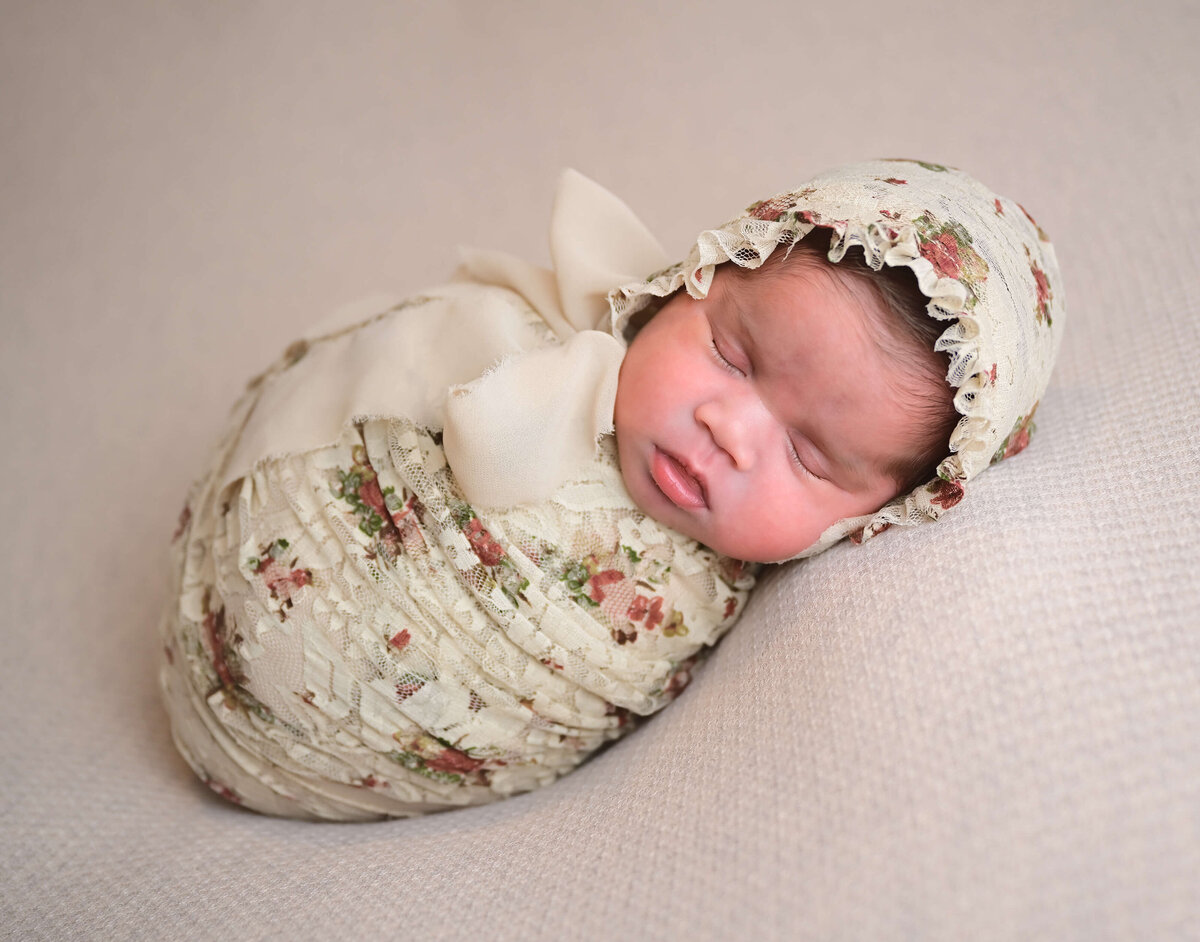 Newborn photo session with baby wrapped in tan floral wrap and matching bonnet on a matching tan blanket. Image by Beth Benecki.