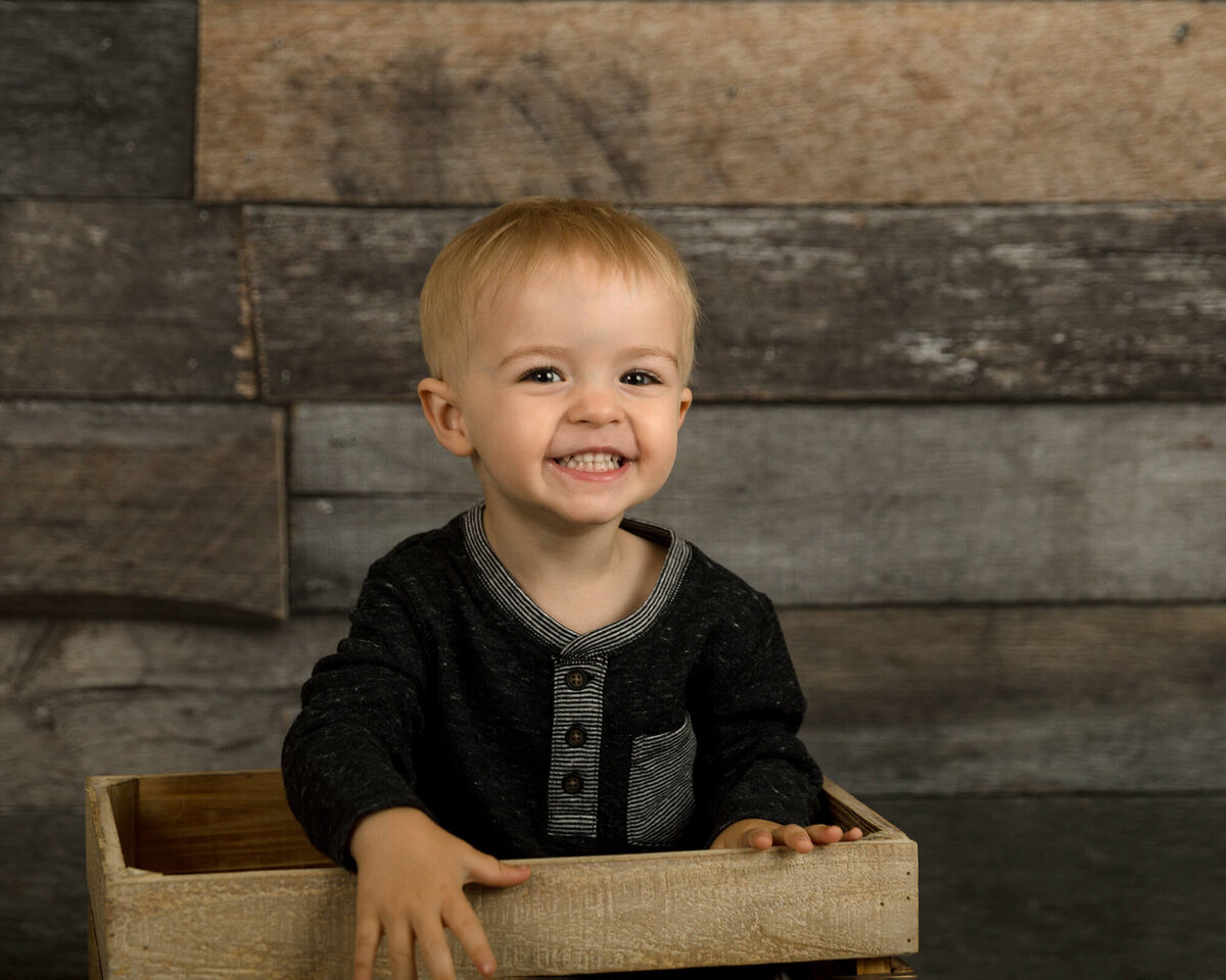 Toddler boy sitting inside a wooden crate with a big smile.