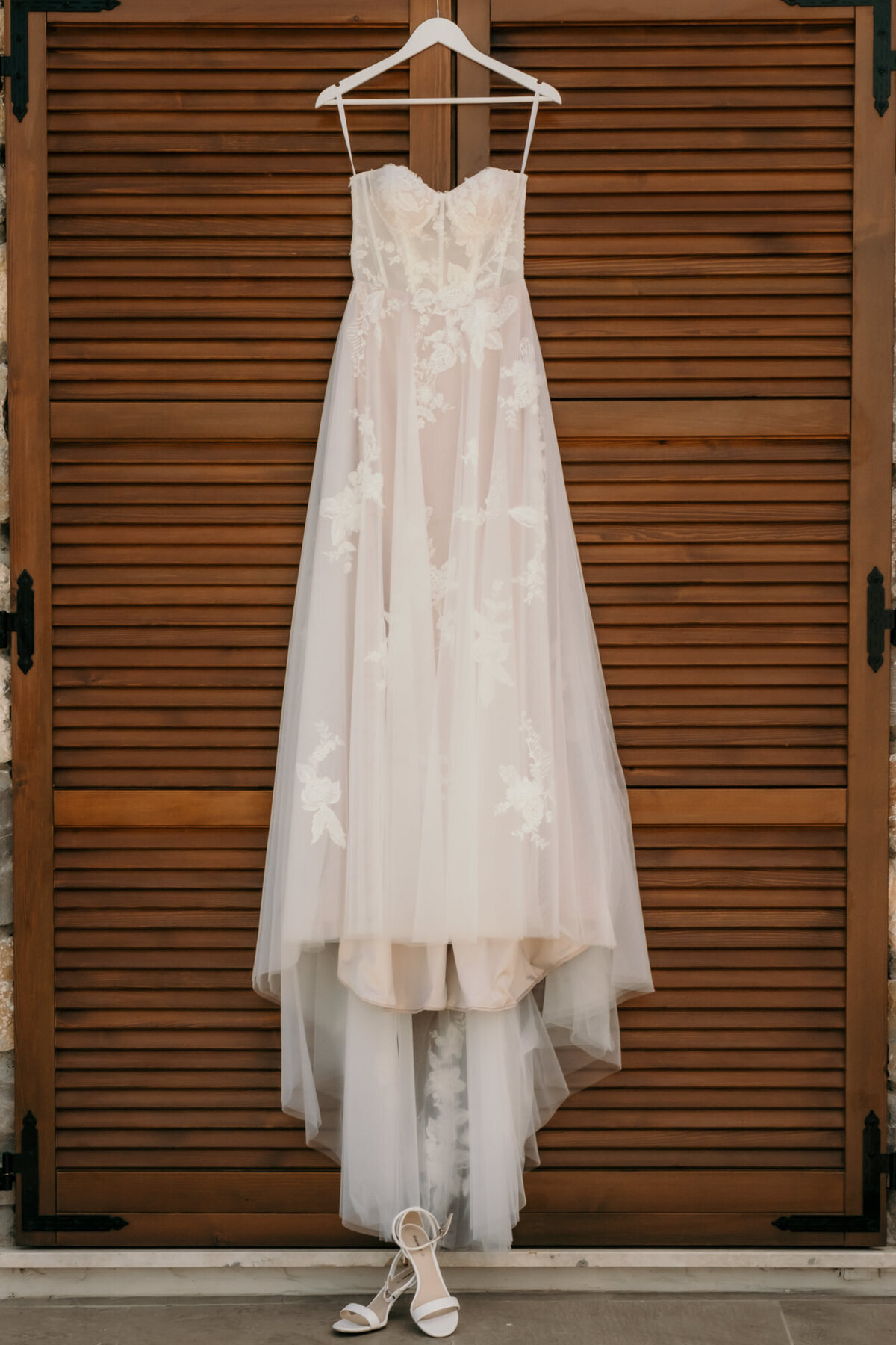 Das trägerlose Brautkleid hängt vor einem Holzfensterladen. Am Boden stehen die Brautschuhe.