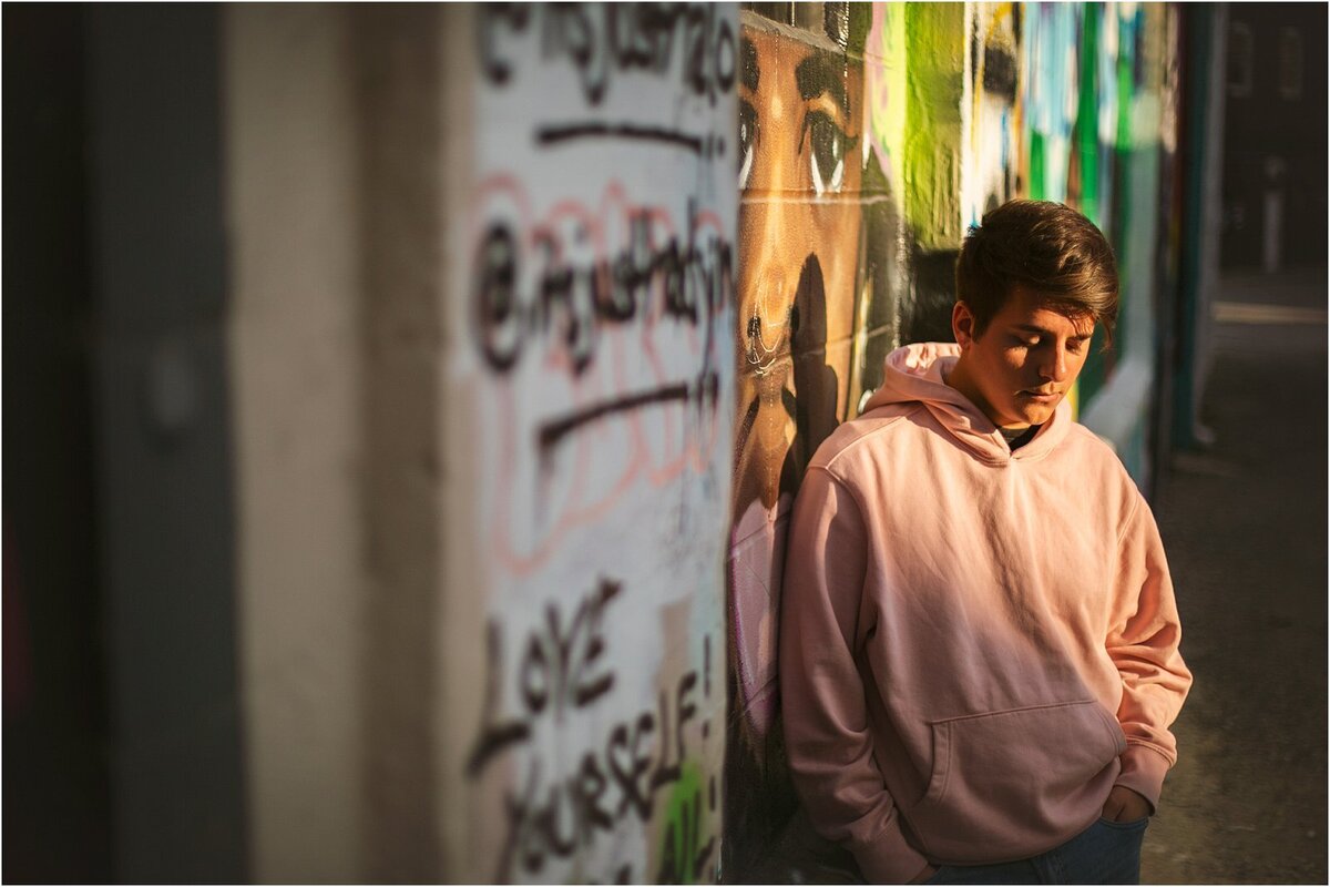 a-high-school-senior-boy-in-a-light-pink-hoodie-leaning-against-a-graffiti-covered-wall-in-an-alleyway-with-dramatic-lighting-during-his-indianapolis-senior-portrait-session-in-fountain-square-indiana