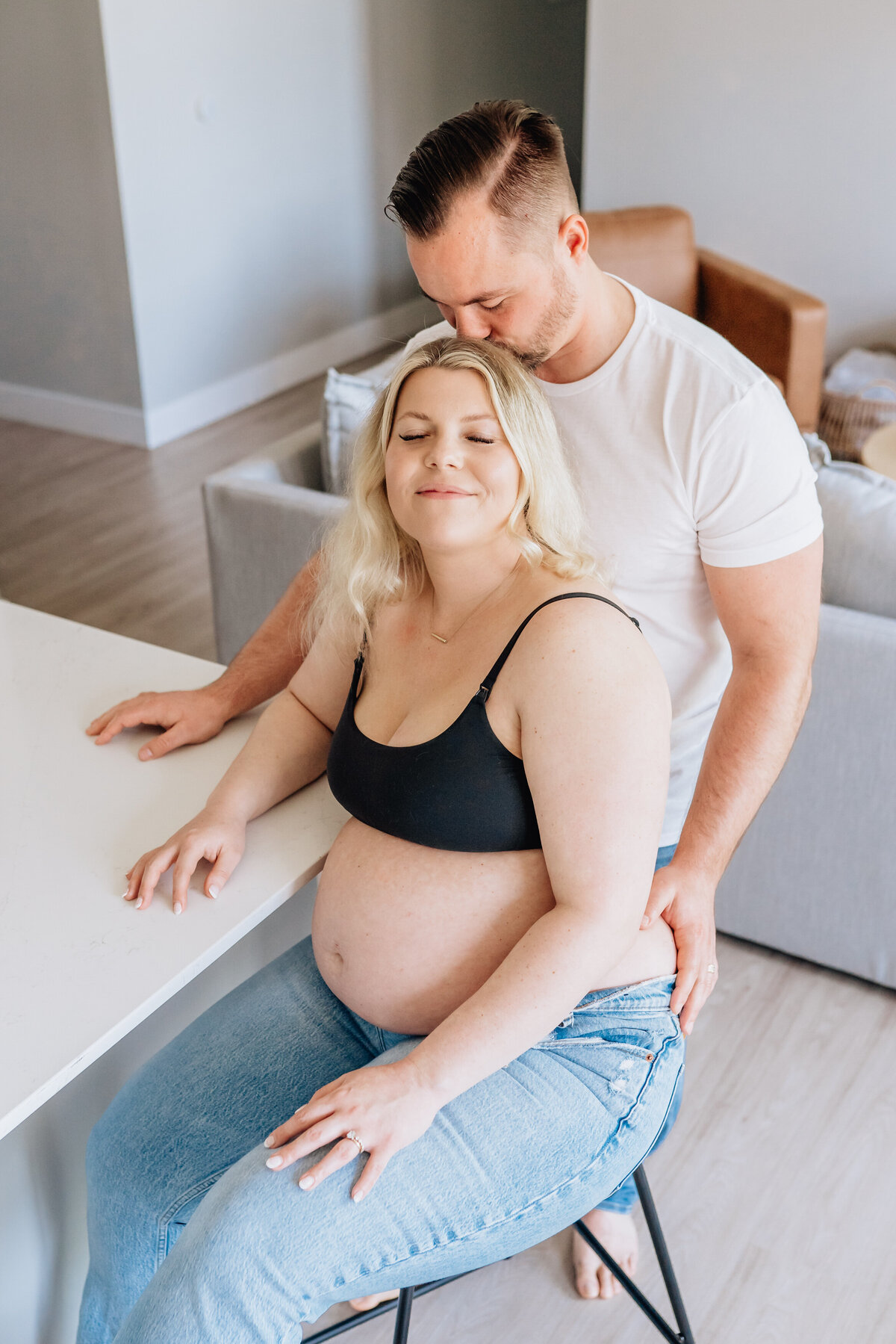 A pregnant woman in a black bra and jeans sits on a stool at a kitchen island, leaning back into her partner who is standing behind her, kissing the top of her head.