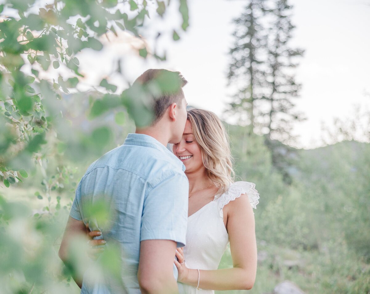 A man in a blue shirt snuggles and kisses his smiling wife in a white dress behind a tree 