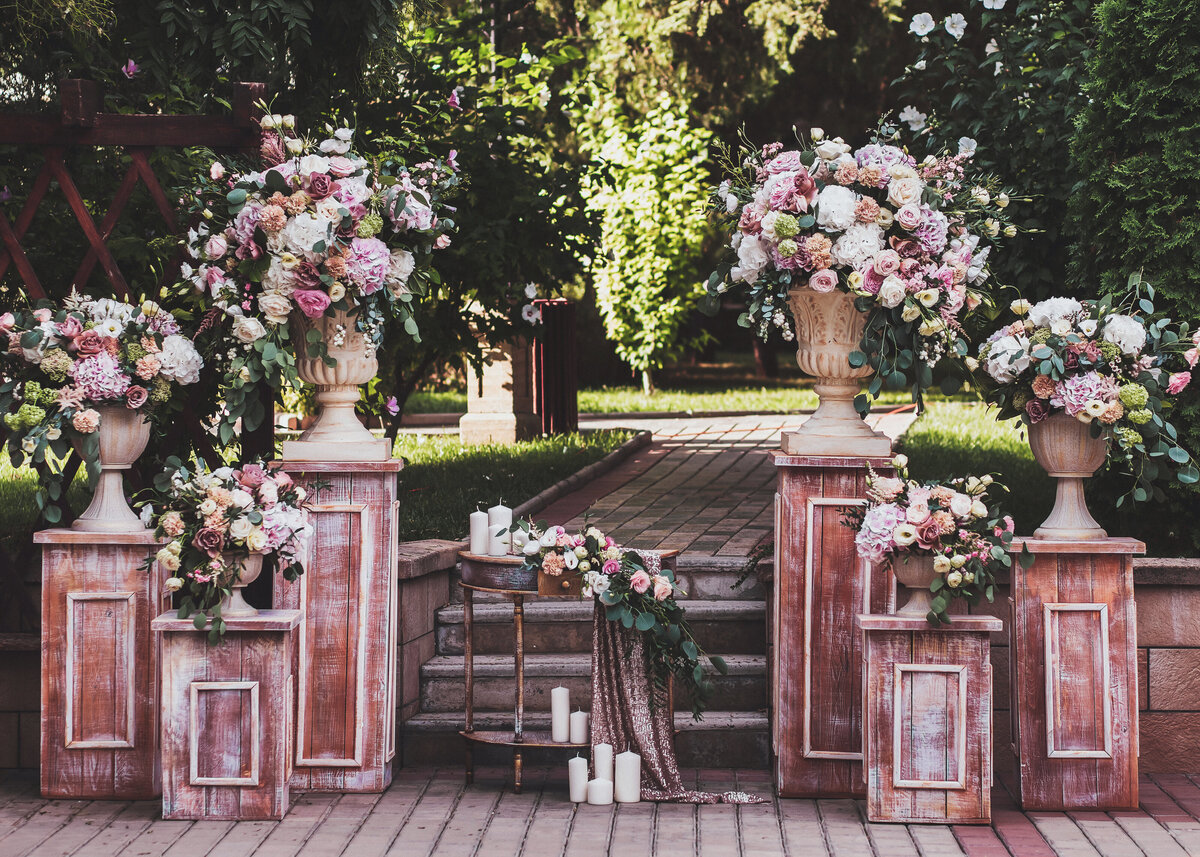 An abundance of roses and hydrangeas are styled in stone urns on wooden plinths as wedding or event decor.