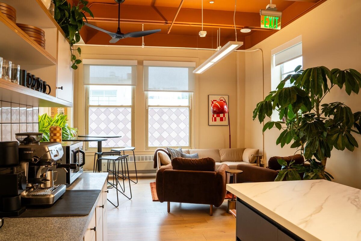 Cozy office lounge and kitchenette featuring warm tan walls, brown velvet seating, a marble island, indoor plants, and a bright orange ceiling with exposed ducts.