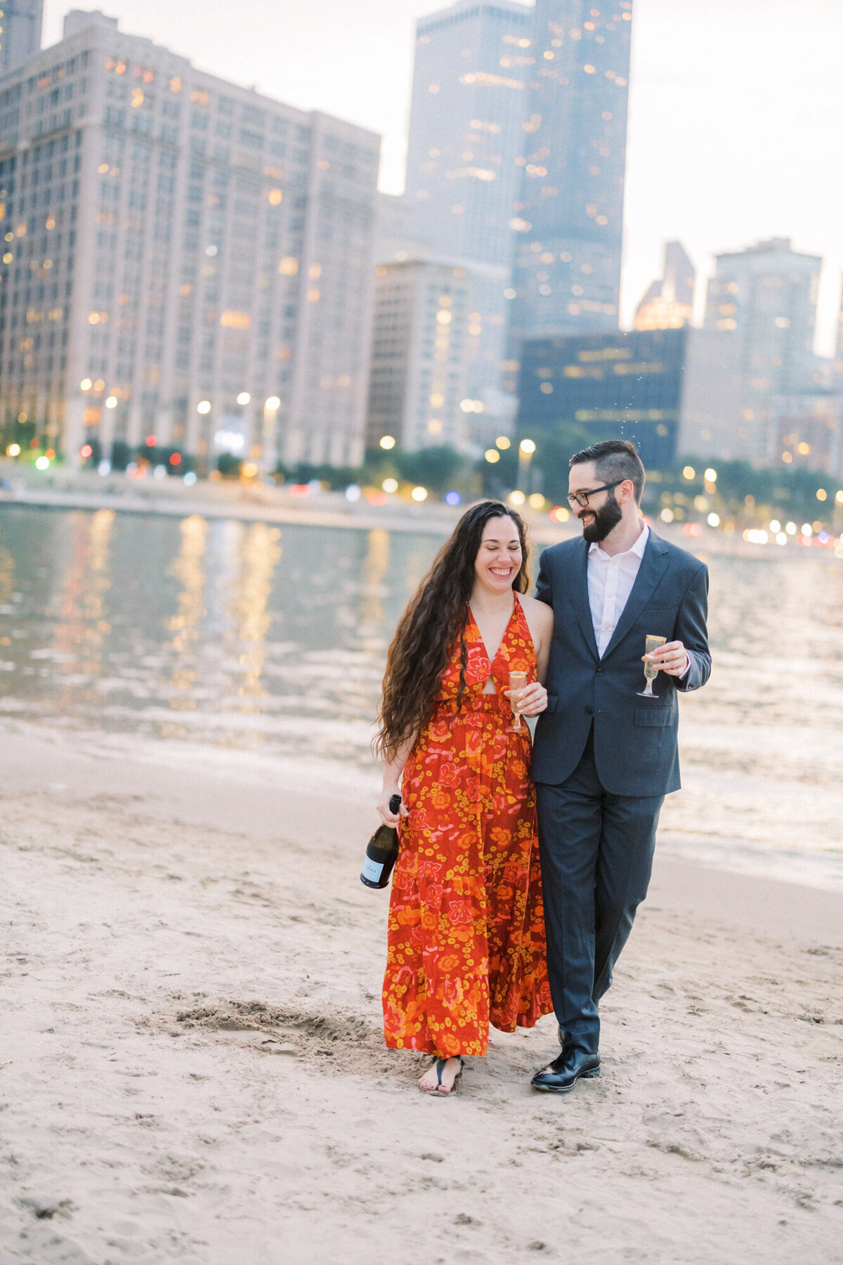 A couple enjoys champagne as they walk along the beach for their engagement photo
