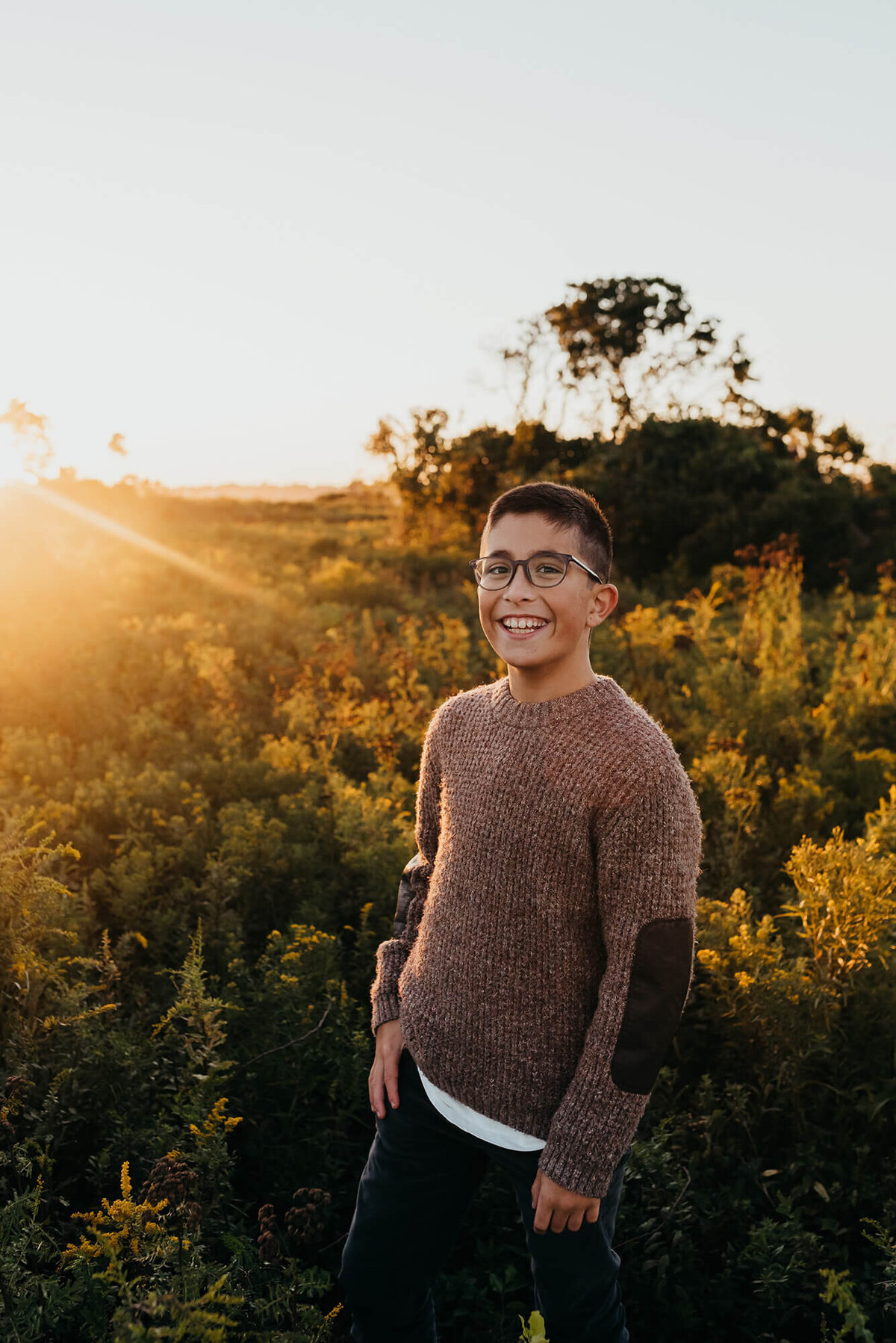 A preteen boy wearing a brown sweater and glasses stands in a field of goldenrod at sunset