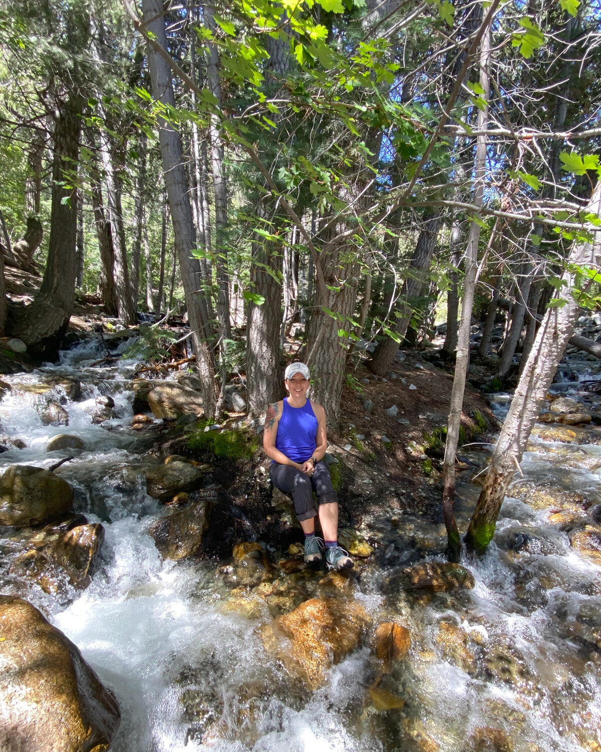 woman sitting under trees with water flowing around her, wearing hiking hear, smiling at the camera