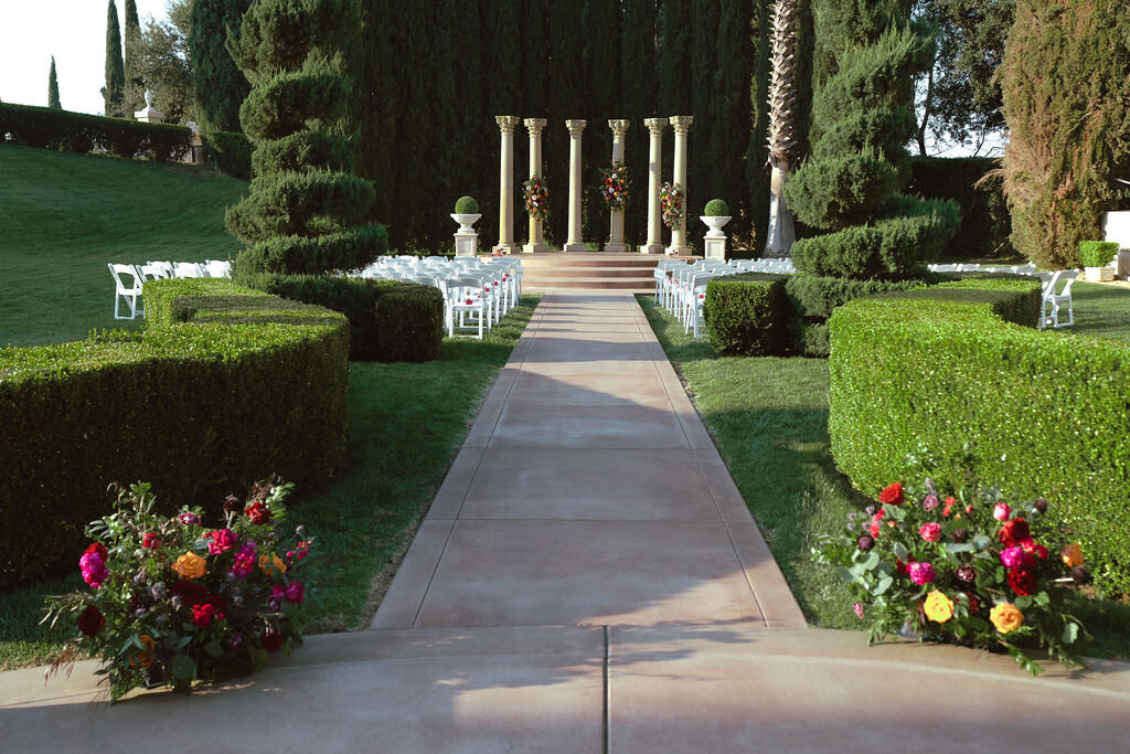 A pathway leading to a setting for a wedding ceremony with colorful flowers and white chairs.
