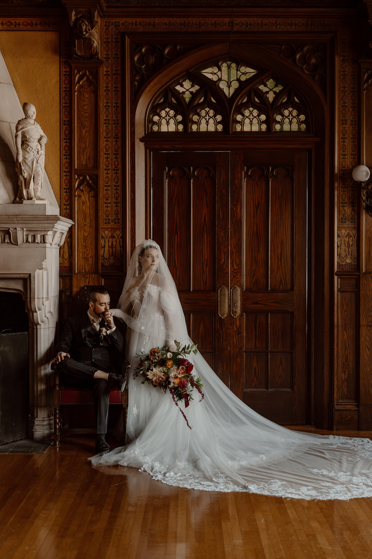 Bride and groom in a historic, elegant room. The bride, dressed in a flowing white gown with a long veil and holding a bouquet of colorful flowers, stands gracefully next to her seated groom. The groom, in a black suit, kisses the bride's hand, adding a romantic touch to the scene.