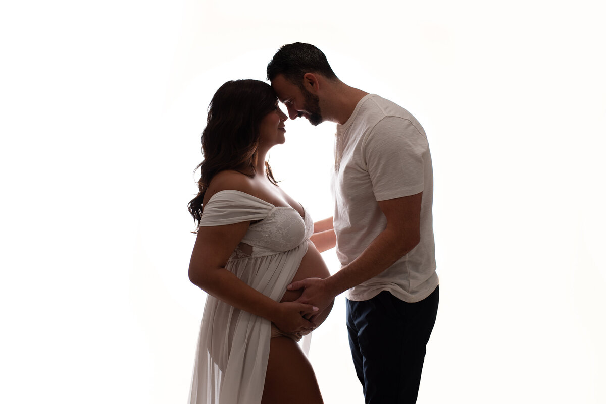 Pregnant woman at maternity and newborn studio of Atlanta posing for maternity portrait wearing white sheer maternity gown head to head with husband holding baby bump
