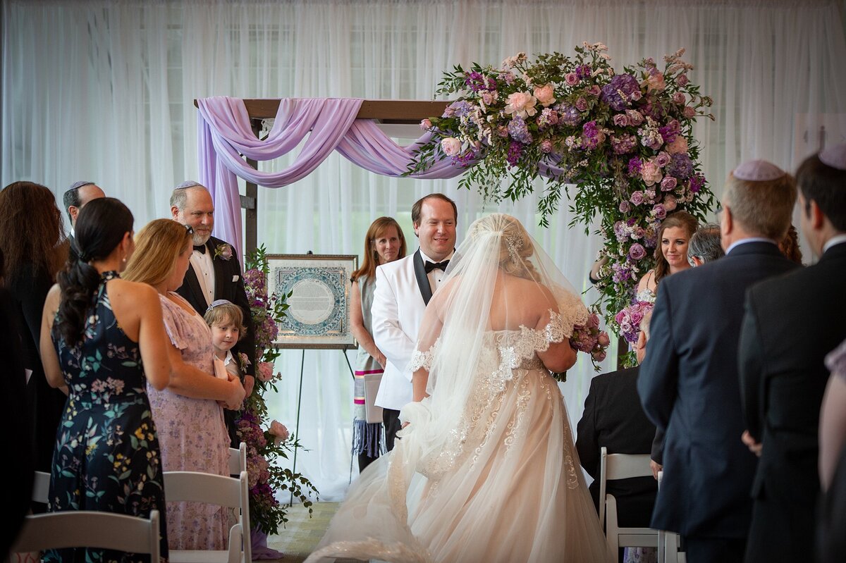 The bride wearing a strapless lace wedding dress and long bridal veil walks the seven steps around the groom who is wearing a white tuxedo with a black shawl collar under the chuppah at The Liff Center in Nashville. The wood chuppah is draped with sheer purple fabric and is decorated with an extra large spray of purple roses, pink roses and greenery.