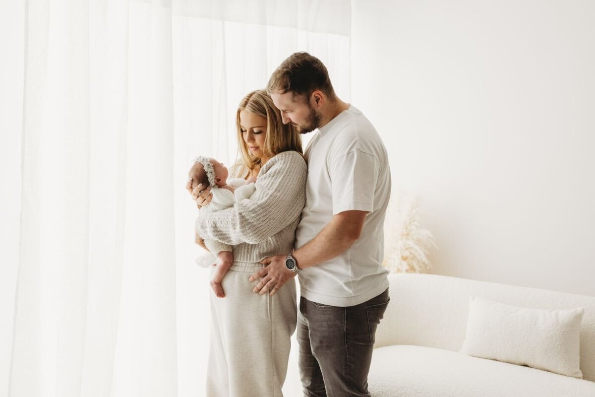a mother and father lovingly holding their peacefully sleeping newborn baby near a bright window by Ruaa Hamdan Photography in UK