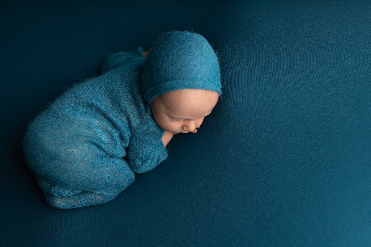 Overhead view of a newborn baby in a teal knit romper peacefully sleeping on teal fabric – Colleyville baby photographer.
