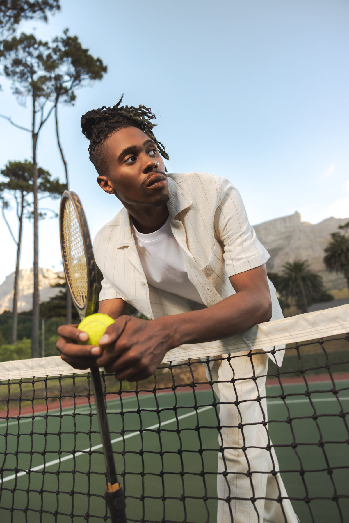 Man leaning over a tennis net holding a yellow ball and racket, dressed in a light summer outfit, with Table Mountain and tall trees in the background under a clear sky in Cape Town.