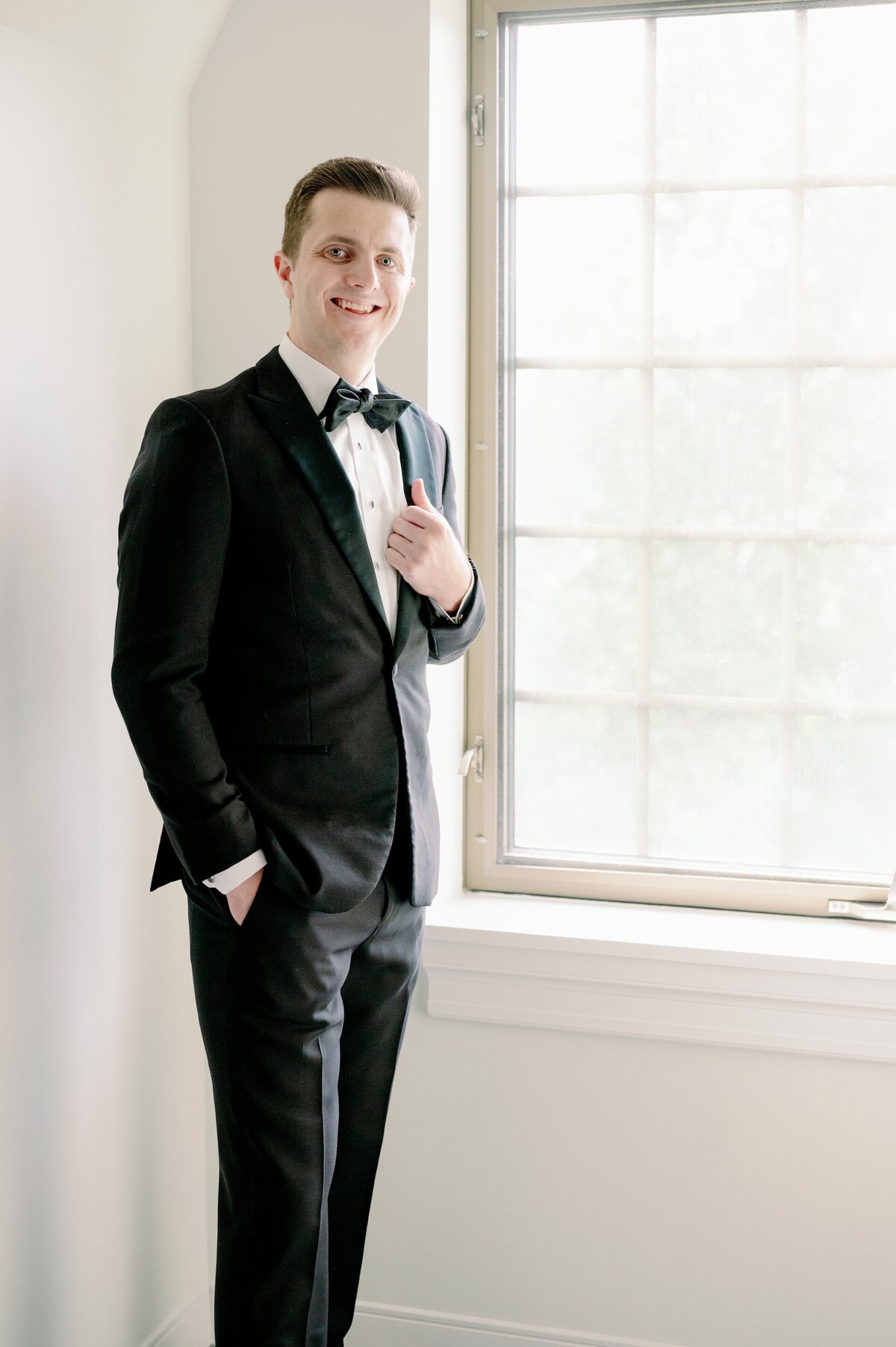 a groom in a tux standing by a window