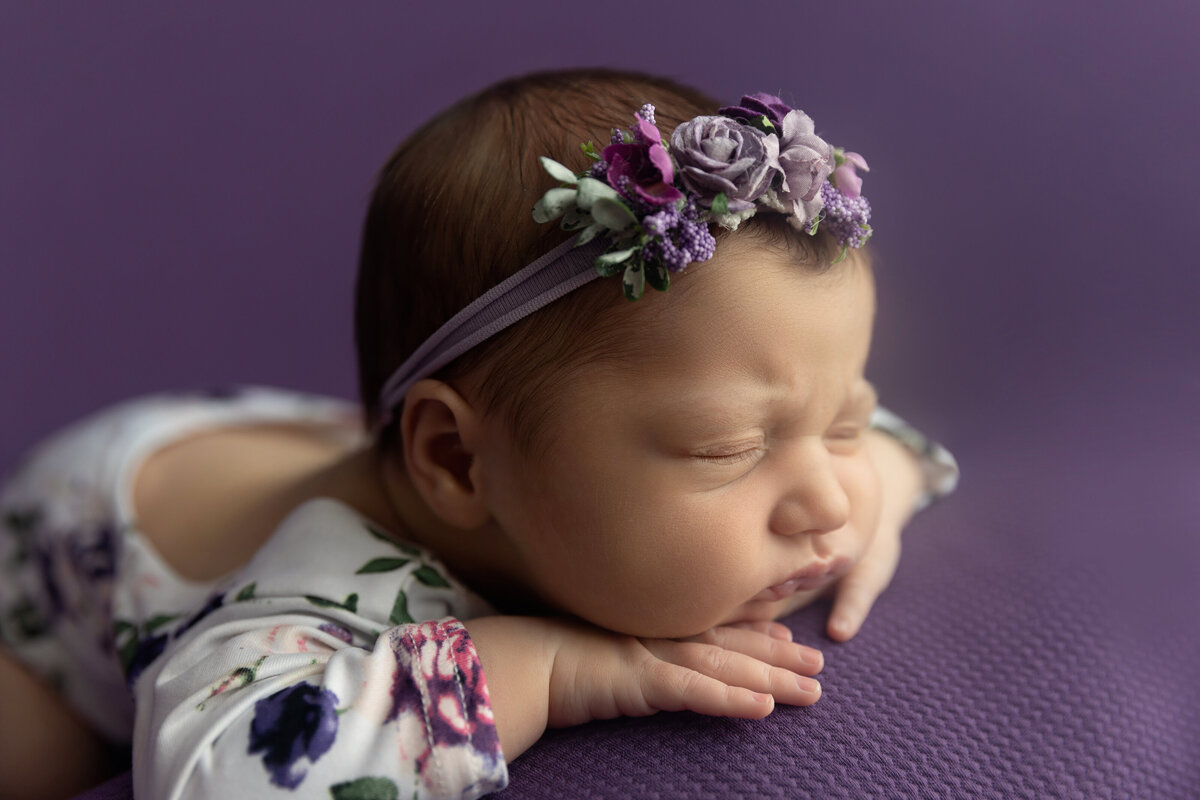 A newborn baby girl in a white and purple floral print floral onesie and matching headband sleeps on her hands on a purple bed posed by a NJ Newborn Photography