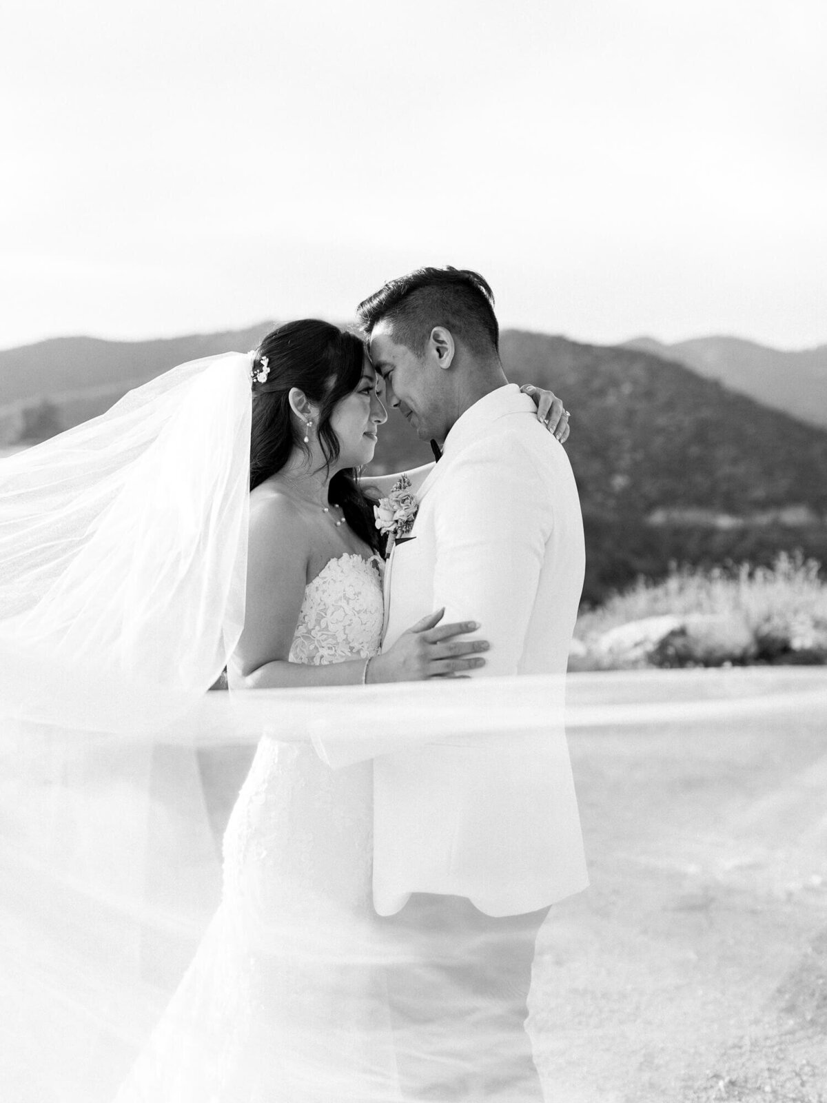 A bride and groom, dressed elegantly, embrace tenderly with foreheads touching. Her flowing veil creates a sense of movement. Mountains form the backdrop.