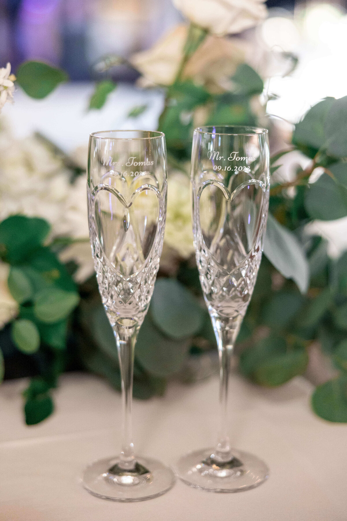 Two crystal champagne flutes engraved with Mr. and Mrs., along with a date, stand elegantly on a table. They are surrounded by lush green foliage and white flowers in the background.