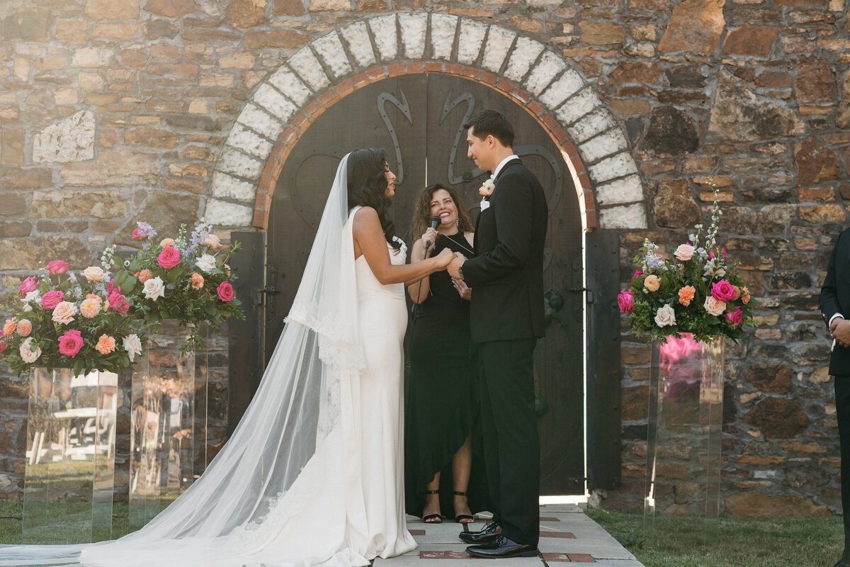 Intimate ceremony moment as the couple exchanges vows in front of a rustic stone chapel, framed by tall floral pedestals overflowing with roses, dahlias, and greenery in pink, peach, and ivory tones.
