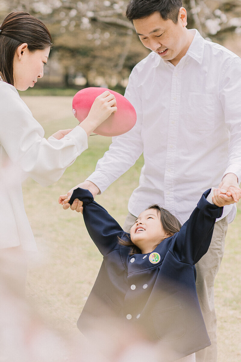帽子をかぶりたくなくて渋っている女の子の手を引いているパパと、かぶせようと帽子を持っているママが笑っている可愛らしい家族写真。相模原市で撮影。