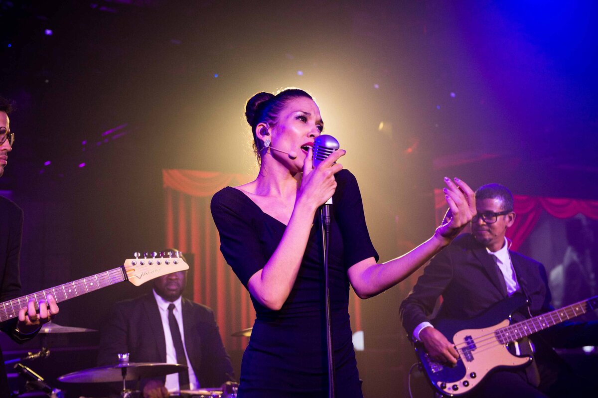 A women holds the microphone to her face in smoky concert hall with band members behind her. A stage light illuminates her from behind for visually stunning composition