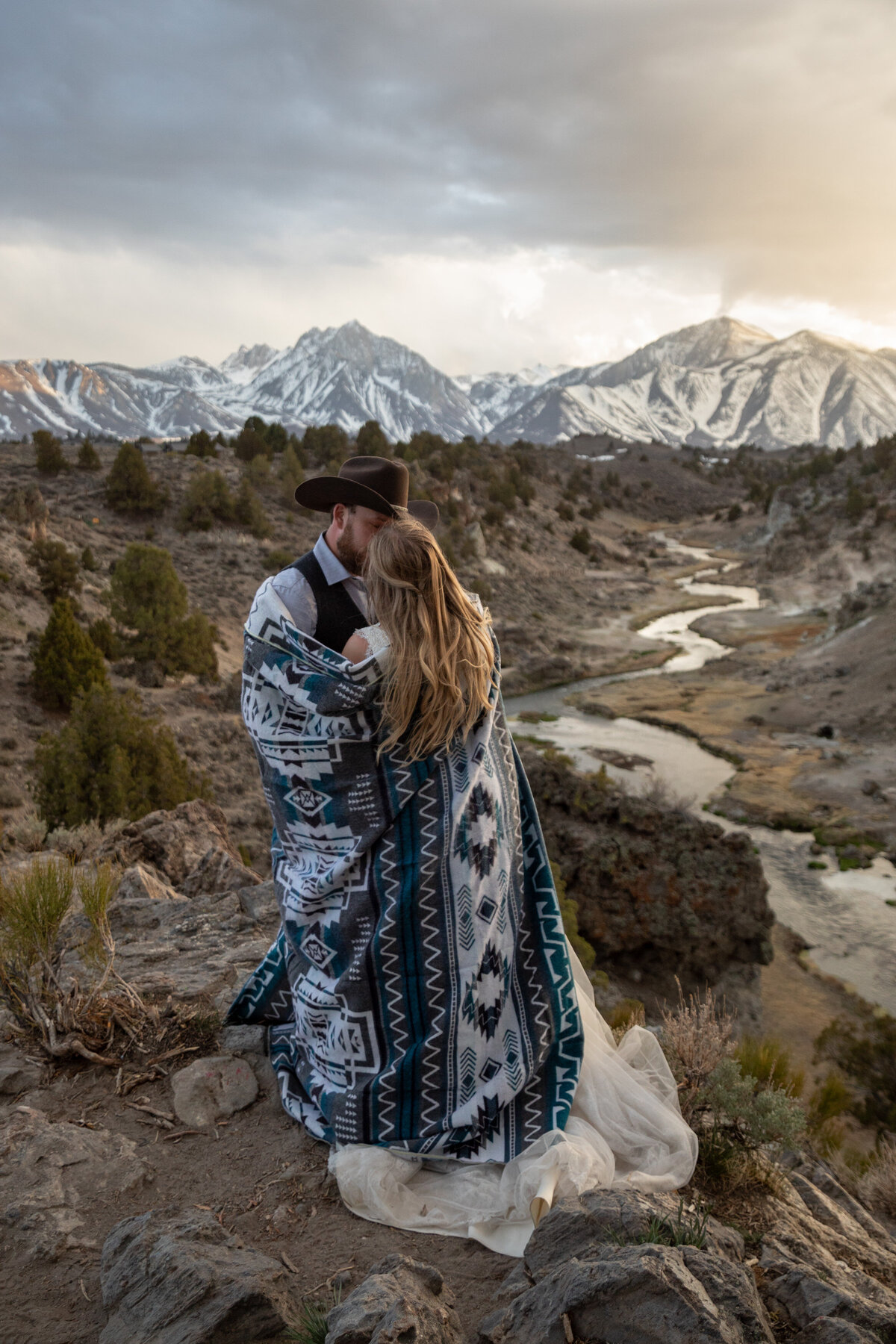A bride and groom stand cuddled up together under a blanket as they watch the sunset over the mountains in the Eastern Sierra.