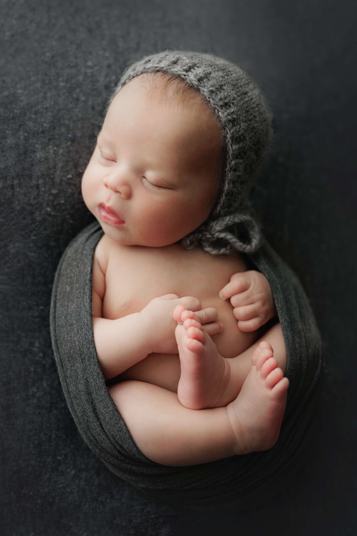 newborn baby laying on his  back with a grey wrap and bonnet at        a photosoot