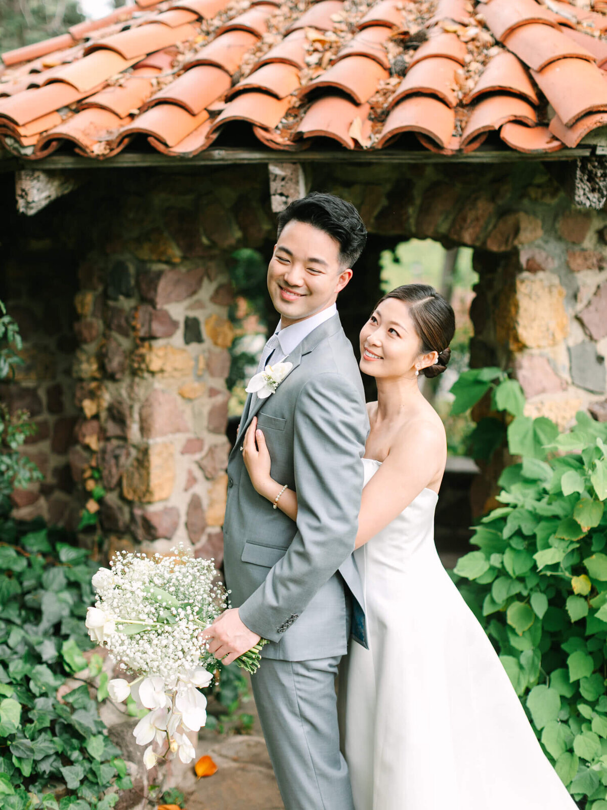 A smiling bride in a white dress embraces the groom in a gray suit, holding a bouquet. They stand in front of a rustic stone wall with lush greenery.