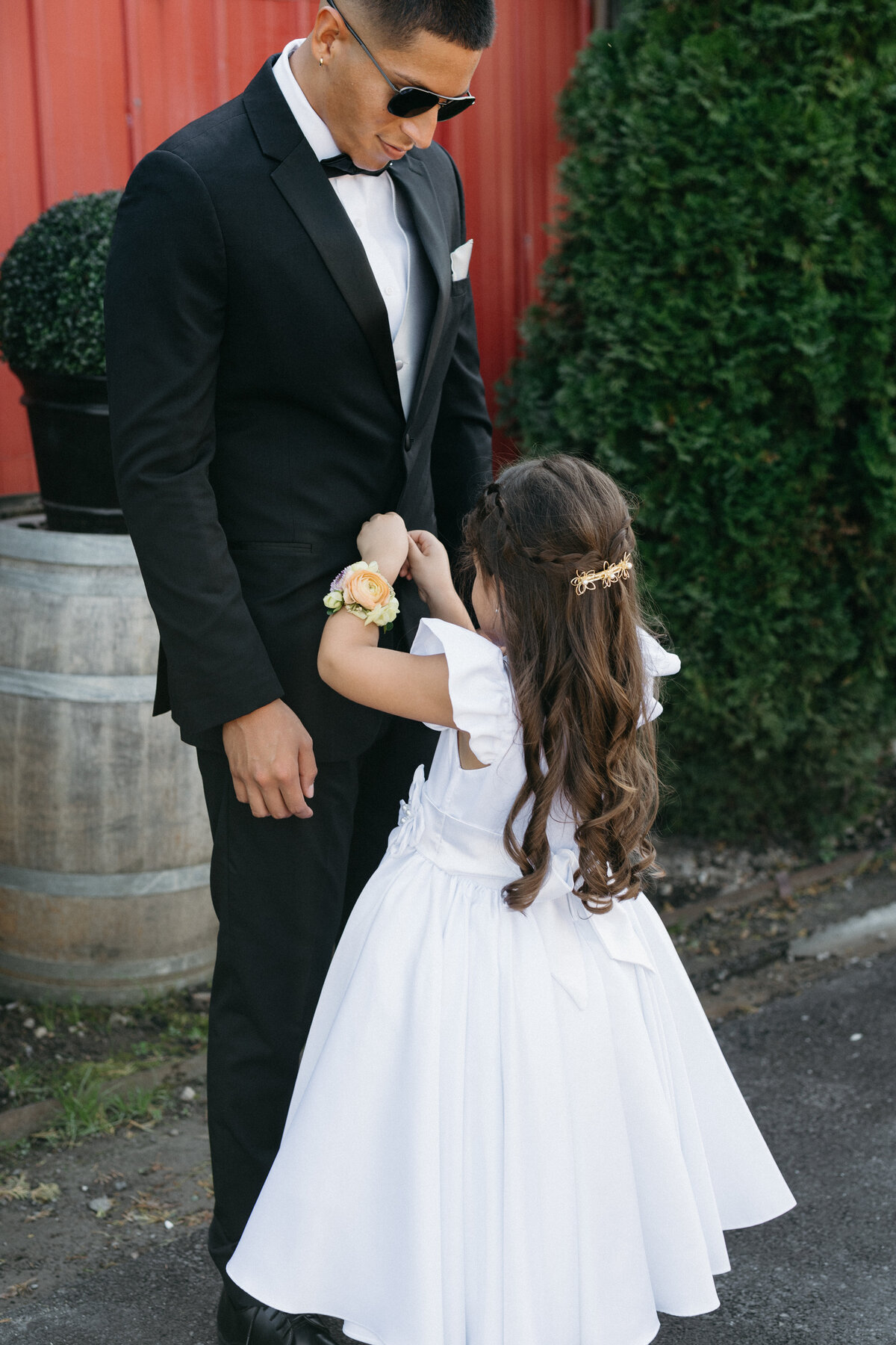 Sweet candid moment of a flower girl in a white dress adjusting the suit of a groomsman while wearing a peach floral wrist corsage.
