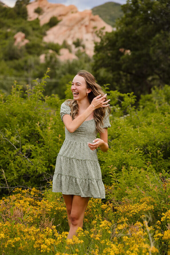 Girl laughs and fixes hair with green bushes and yellow flowers behind her.
