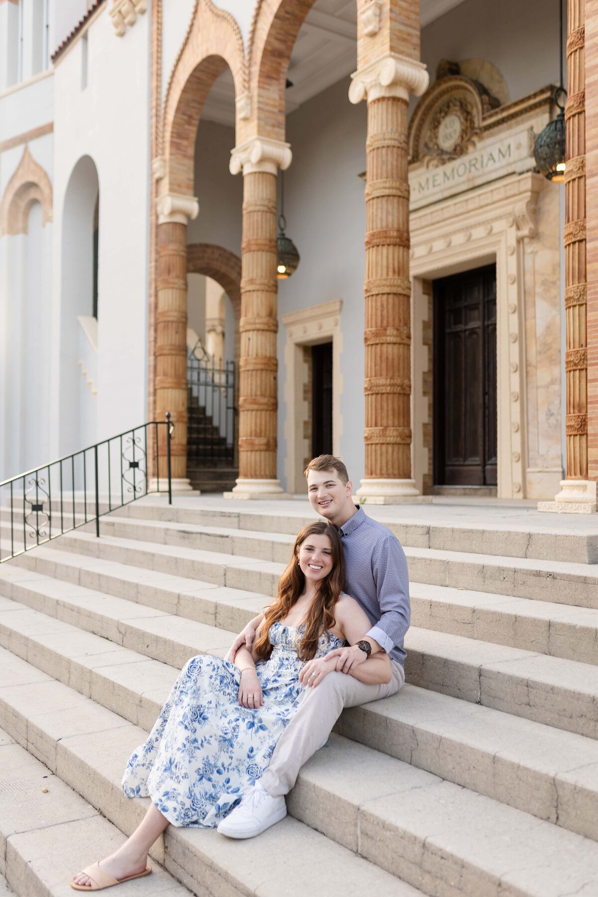 A couple sits together on the grand steps of a historic building in St. Augustine, Florida, surrounded by detailed stone columns and elegant architecture. The bride-to-be wears a flowy blue and white floral dress while resting against her fiancé, who wears a light purple button-down shirt. Their relaxed pose and bright smiles capture the excitement of their engagement. Photographed by Heartful Films, a Jacksonville engagement photographer and videographer known for timeless imagery in romantic, architectural settings.