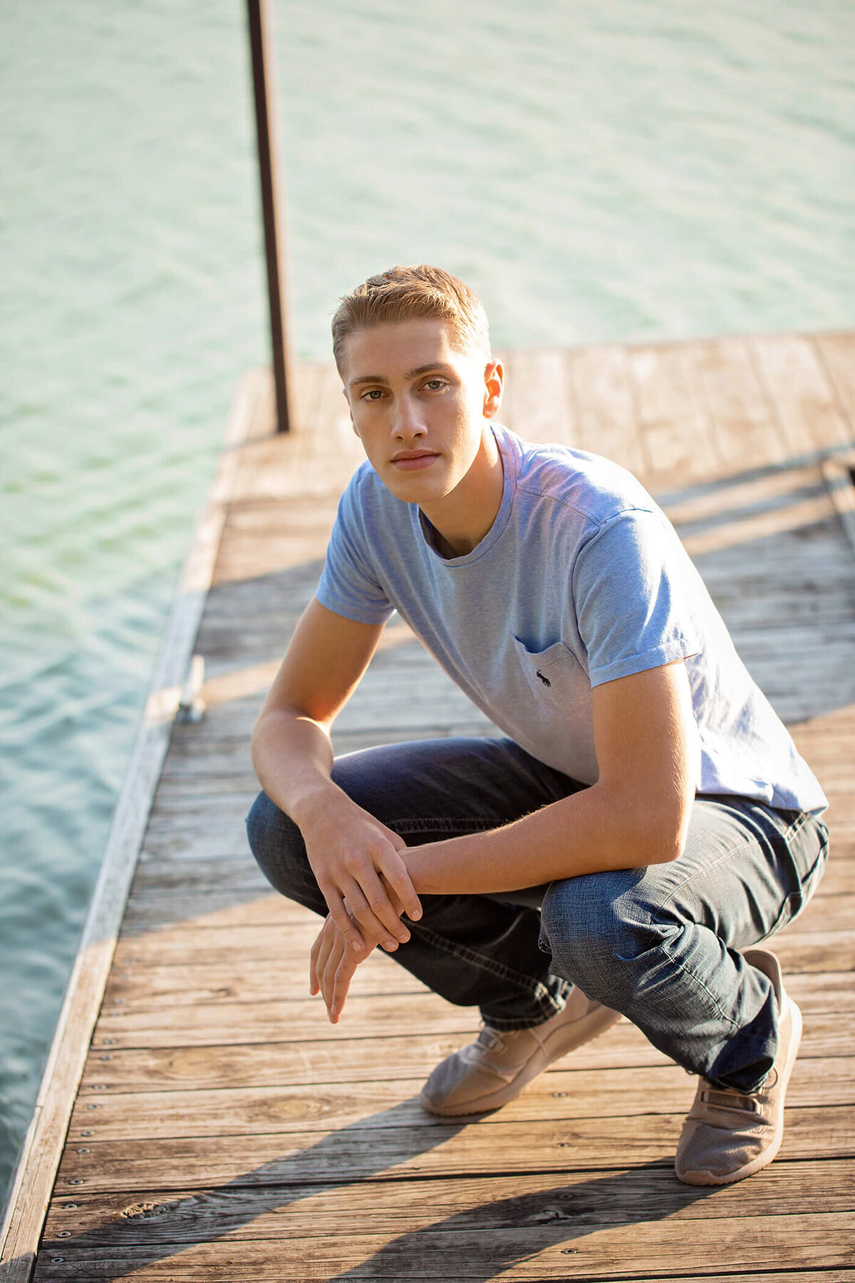 Senior boy squatting down at the end of a pier with water surrounding.