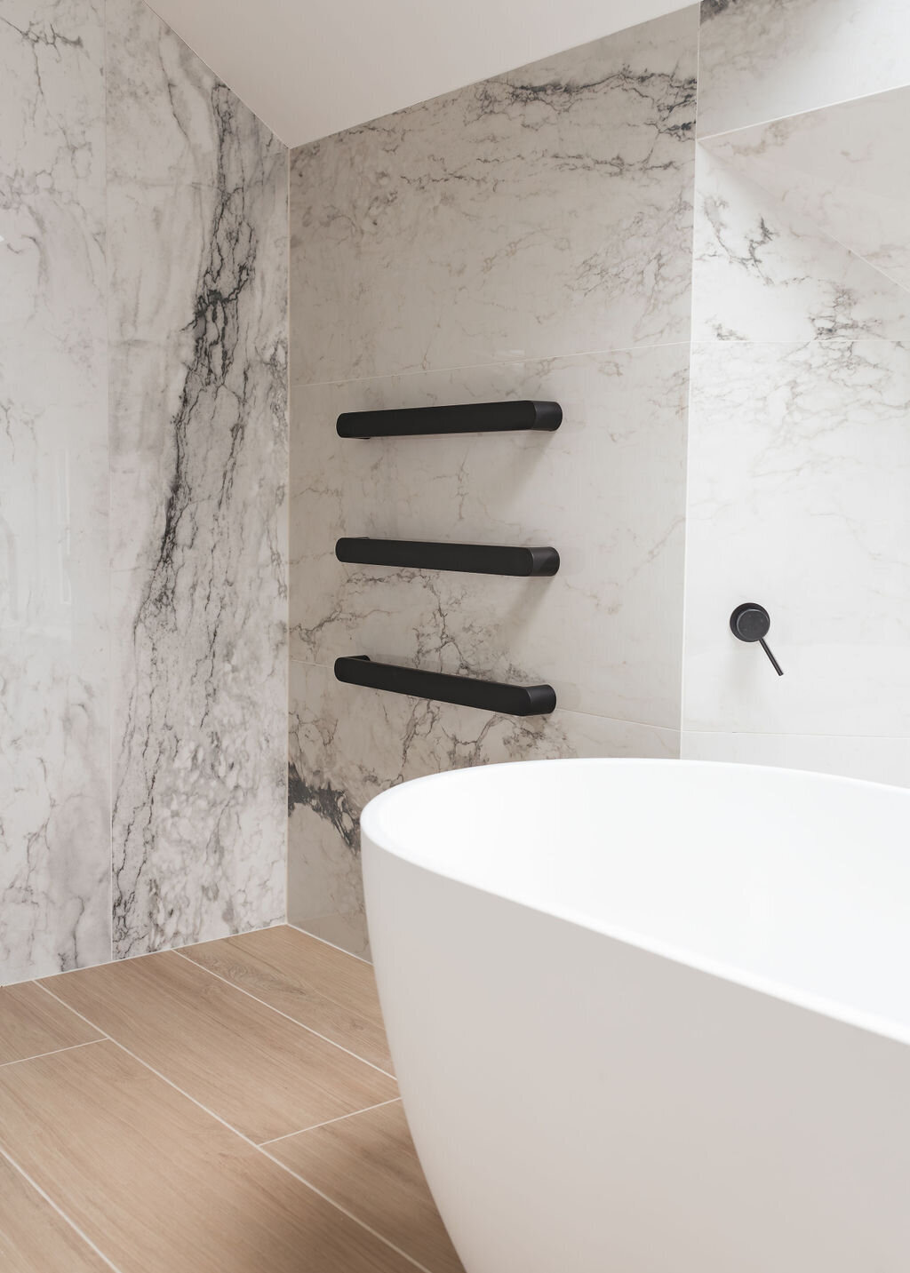 A modern white soaking tub in a grey marble bathroom with elegant black towel bars mounted to the marble walls.